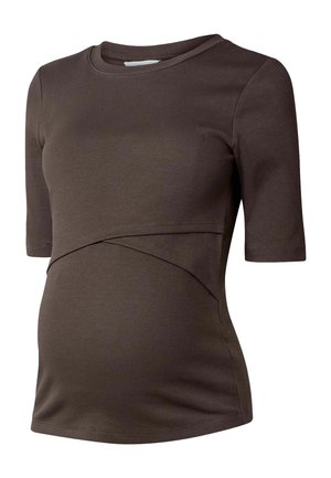 Femme enceinte portant une tenue brune ajustée et une veste en cuir brun foncé drapée sur ses épaules, sur un fond beige uni.