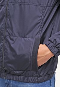 Giacca leggera navy in tessuto morbido con chiusura a zip, tasche laterali e polsini elasticizzati. Presenta dettagli di cucitura lungo le cuciture.