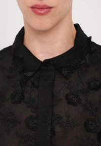 Gros plan d'une personne portant un chemisier noir transparent avec broderie florale et col boutonné, ne montrant que les lèvres et le cou.