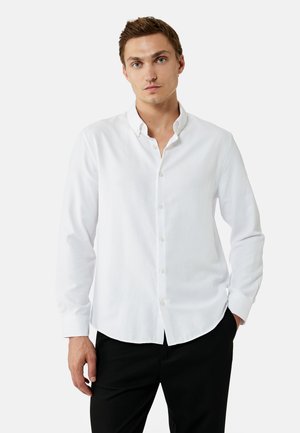 Junger Mann steht mit einer Hand in der Hosentasche, trägt ein weißes Hemd mit Knöpfen und schwarze Hosen vor einem schlichten weißen Hintergrund.