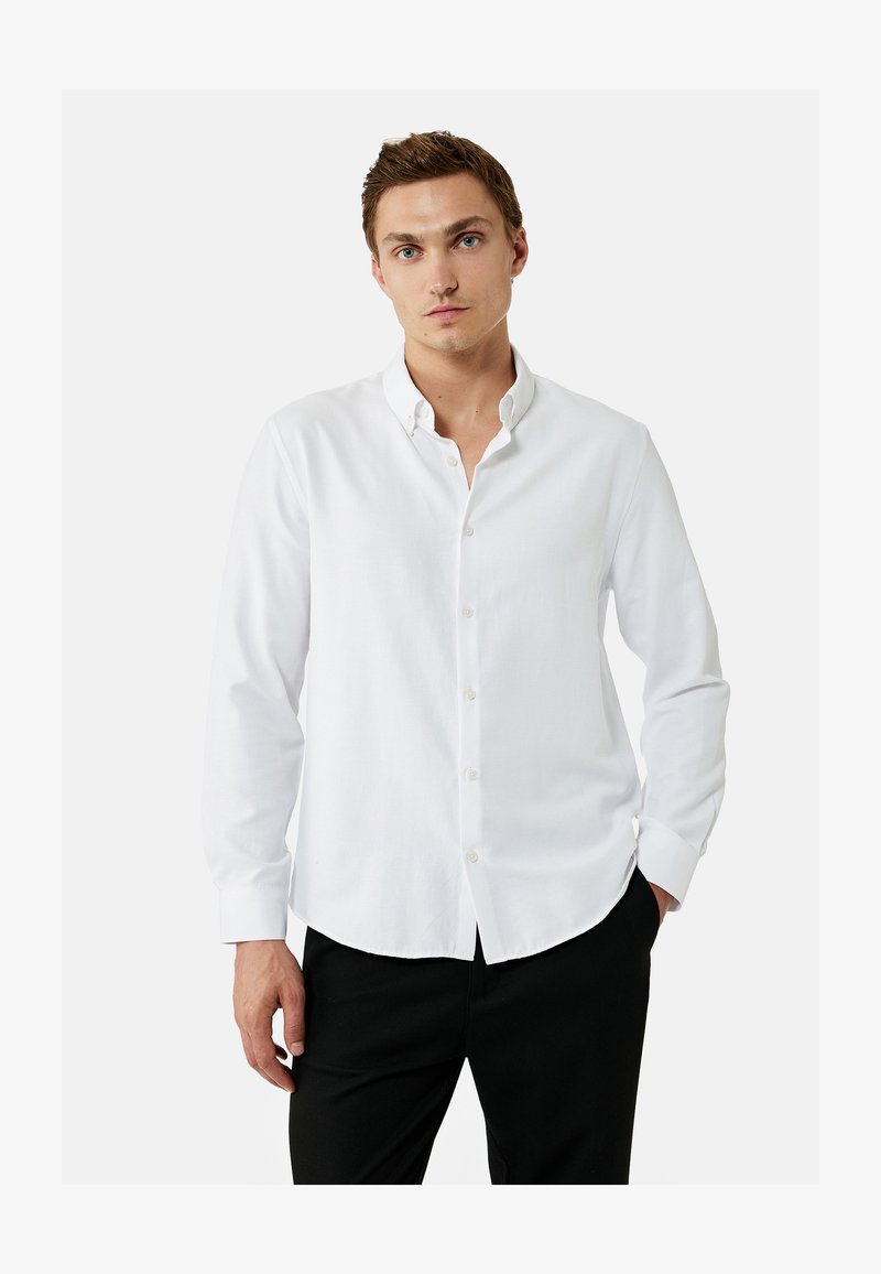 Young man standing with one hand in pocket, wearing a white buttoned shirt and black pants against a plain white background.