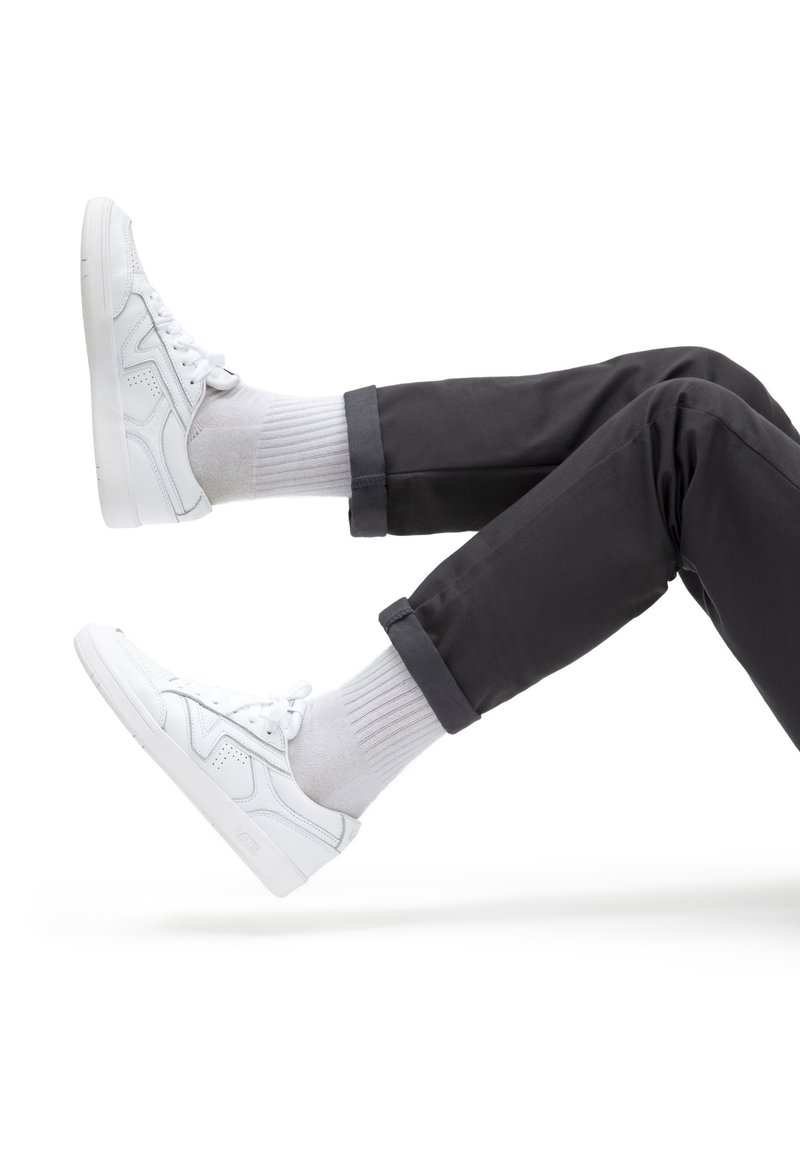 Personne portant un pantalon noir, des chaussettes blanches et des baskets blanches à lacets, jambes pliées aux genoux sur fond blanc.