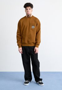 Sudadera marrón de mezcla de algodón, con un bolsillo que presenta un detalle del logo en negro. Combinada con pantalones holgados negros y zapatillas negras.