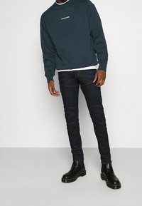 Sweatshirt vert foncé avec logo blanc, superposé à une chemise blanche. Jean slim-fit bleu foncé avec coutures visibles, associé à des bottes noires.