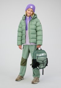 Groene pufferjack, lila tussenlaag, groene broek met donkere accenten, en een groene rugzak met dierenafbeeldingen. Beige schoenen maken de outfit compleet.