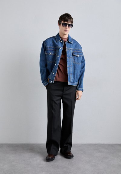 Chaqueta de mezclilla azul con dos bolsillos frontales, llevada sobre una camiseta marrón. Unos pantalones de pierna ancha negros y zapatos oscuros completan el conjunto.