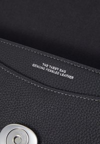 Interno di una borsa in pelle nera con effetto granigliato e hardware di colore argento. Presenta cuciture lungo il bordo e un logo impresso con la scritta "THE TABBY BAG."