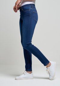 Jeans de mezclilla azul delgados con cintura alta, que cuentan con cinco bolsillos y costuras sutiles. Combinados con zapatillas blancas para crear contraste.
