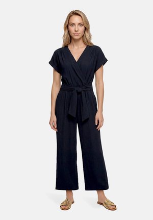 Femme portant une combinaison noire à manches courtes avec une ceinture à la taille et des jambes larges, associée à des sandales plates dorées, debout sur fond blanc.