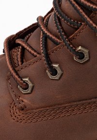 Bota de cuero marrón con acabado texturizado, con cordones negros y marrones, ojales metálicos hexagonales y costuras detalladas a lo largo de las uniones.