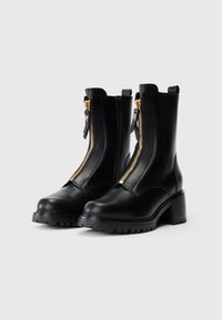 Botines de piel negra con tacones gruesos y cremalleras frontales doradas prominentes sobre un fondo blanco.