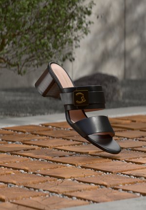 Μαύρο πέδιλο με ανοιχτά δάχτυλα και τακούνι, με δύο λουριά, λεπτομέρεια χρυσής αγκράφας στο πάνω λουρί και χοντρό ξύλινο τακούνι σε πλακόστρωτη εξωτερική επιφάνεια.