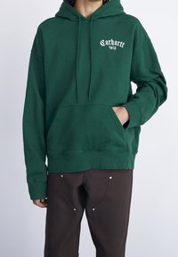 Sudadera verde con capucha y bolsillo canguro, puños y bajo acanalados, con el logo blanco de "Carhartt WIP" en el pecho. Tela suave, ajuste casual.