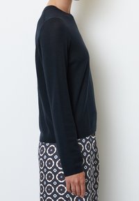 Pull noir à manches longues en matériau doux, coupe ajustée, encolure ronde, porté avec une jupe à motifs géométriques.