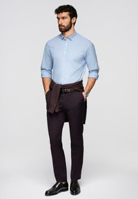 Homme barbu portant une chemise boutonnée bleu clair, un pantalon foncé, des chaussures habillées noires, et un pull foncé noué autour de la taille, debout les mains dans les poches.