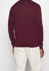 Maroon knit sweater with a crew neck, ribbed cuffs, and hem. Features a small rectangular detail at the back neckline. Soft texture.
