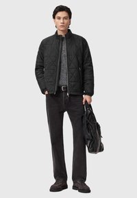 Czarna pikowana kurtka z kołnierzem stojącym, szary sweter z dzianiny, czarne dżinsy straight-leg oraz brązowe buty na sznurówki. Trzyma czarną torbę.