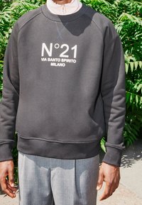 Felpa nera con testo bianco "N°21 VIA SANTO SPIRITO MILANO", dotata di colletto girocollo e polsini a costine, abbinata a pantaloni grigi chiari.