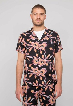 Homme portant une chemise noire à manches courtes avec des boutons et un pantalon imprimé de grandes fleurs roses et pêche, debout devant un fond blanc.