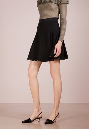 Vrouw draagt een zwarte gerokte rok met kleine studs aan de zoom, een geribd shirt met lange mouwen en zwarte slingback schoenen met lage hak.