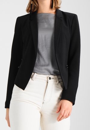 Femme portant un blazer noir ajusté sur un haut gris et un jean blanc taille haute, avec de longues boucles d'oreilles noires à pompons visibles.