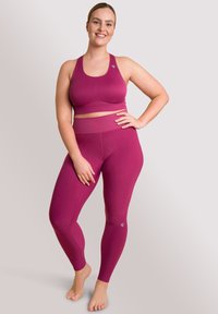 Uśmiechnięta kobieta stoi boso w różowym staniku sportowym i legginsach, z jedną ręką na biodrze, a drugą opartą wzdłuż ciała.