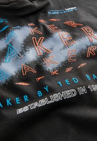 Melna kapucēsceļa ar daudzkrāsainu tekstu "BAKER" zilā un oranžā krāsā, ar tekstūras gradienta efektu. Zem tā, "ESTABLISHED IN 1987."