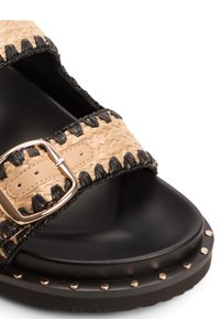 Woven natural straw and black fabric sandal with a gold buckle, flat black sole, and metal studs along the edge.