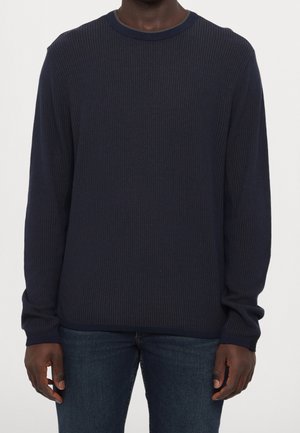 Dorosły ubrany w granatowy, prążkowany sweter z długim rękawem, zestawiony z niebieskimi jeansami o średnim odcieniu, stojący na jednolitym tle.