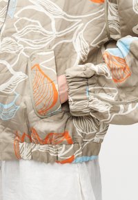 Μπεζ καπιτονέ μπουφάν με λευκά, πορτοκαλί και μπλε λουλουδένια σχέδια, ελαστικά μανσέτες και τσέπη με καπάκι που έχει ένα χέρι μέσα.