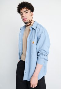 Vaaleansininen nappikauluspaita, jossa on käännetyt hihat ja rintatasku, jossa on oranssi logo, päällä beige crewneck. Mustat housut.