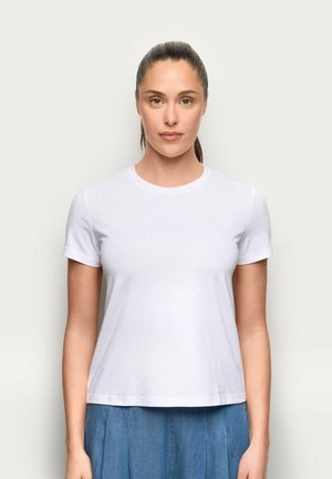 Kobieta z zaczesanymi do tyłu włosami, ubrana w prostą białą koszulkę z krótkim rękawem i niebieską plisowaną spódnicę, stojąca przodem na jasnym tle.