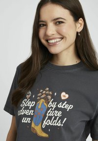 T-shirt gris avec un design graphique d'une botte de cowboy ornée de fleurs, texte "Pas à pas" en blanc et orange, encolure ronde, en coton.