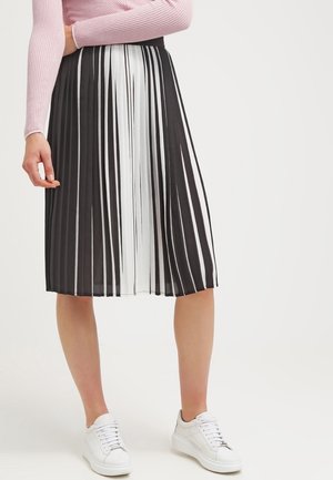 Mujer con falda midi plisada de rayas verticales en blanco y negro, camiseta de manga larga rosa y zapatillas blancas, de pie frente a un fondo blanco.