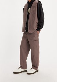 Světle hnědá bezrukávová vesta přes černou mikinu s kapucí, spárovaná se světle hnědými cargo kalhotami; bílé boty s černými tkaničkami a silnými podrážkami.