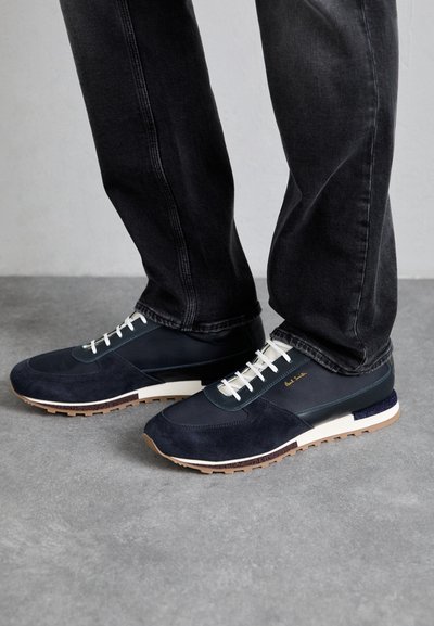 Navy blue sneakers made of suede and leather with white laces, rubber sole, and contrasting accents. Worn with dark jeans.