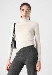Bež rolka od mekog materijala, u kombinaciji s tamnosivim vunenim hlačama. Model drži malu crnu torbicu.