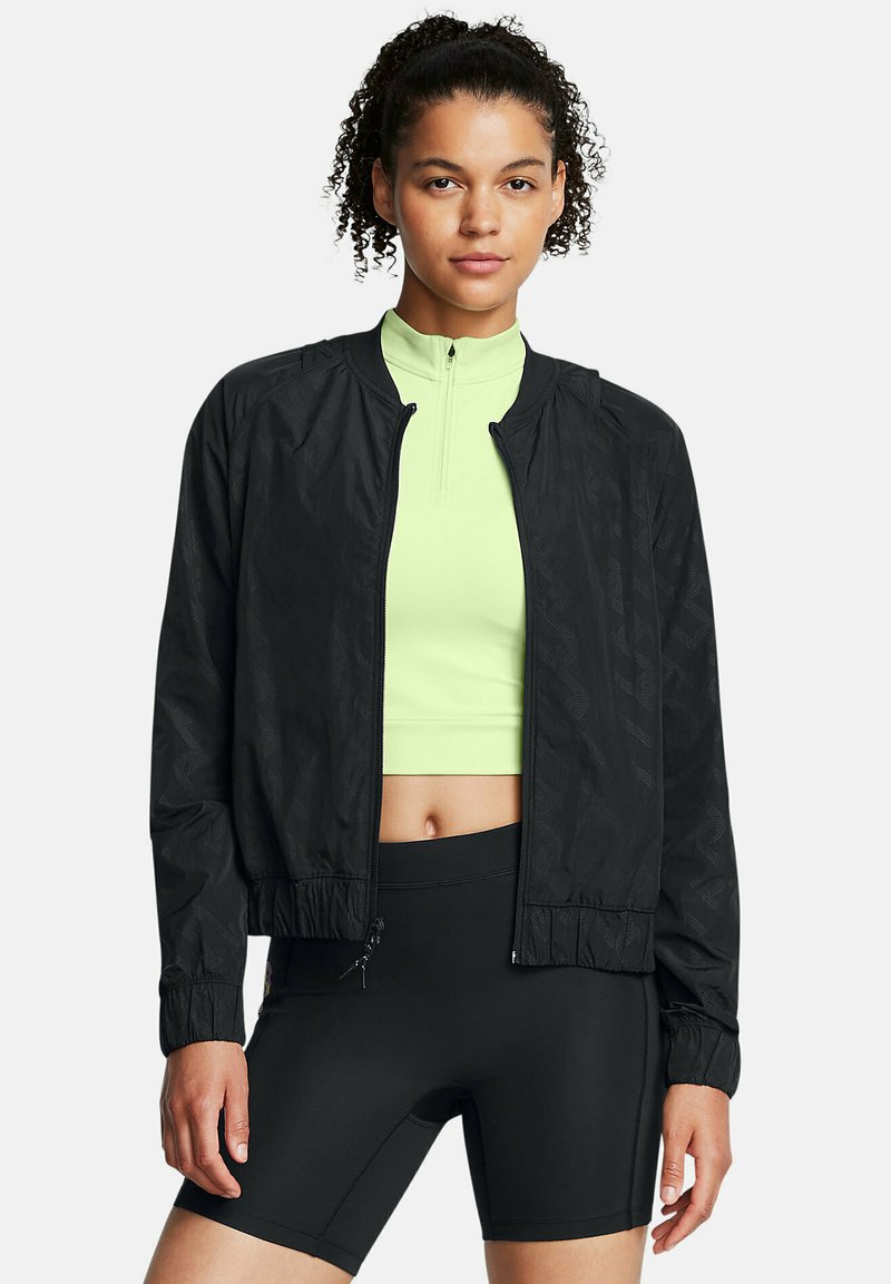 Schwarze Bomberjacke mit subtiler Textur, Reißverschluss und elastischen Bündchen. Getragen über einem hellgrünen, figurbetonten Oberteil und schwarzen Shorts.