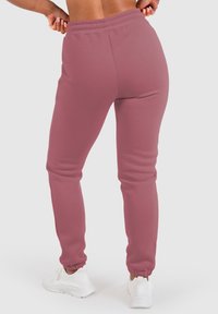 Pantalones de chándal rosas con tobillo ajustado y cintura elástica, hechos de tela suave, usados con zapatillas deportivas blancas. Se muestra la vista desde atrás.