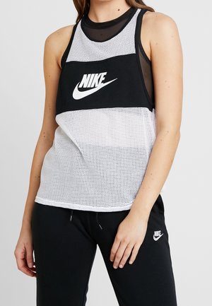 Femme portant un débardeur en maille noir et blanc de Nike et un pantalon de survêtement noir Nike avec logo blanc, debout avec une main près de la taille.