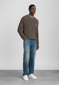 Καφέ ριγέ πουλόβερ με λαιμόκοψη και μικρό λογότυπο, συνδυασμένο με ξεθωριασμένο ίσιο τζιν σε μπλε απόχρωση και λευκά αθλητικά παπούτσια.