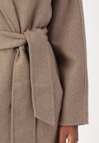 Manteau en laine marron avec une taille ceinturée, doté d'un tissu texturé, de larges revers et de grandes poches latérales.