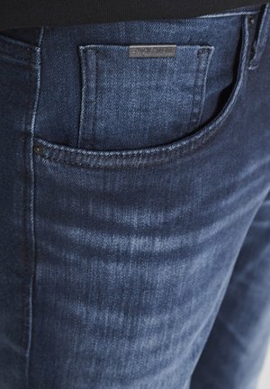 Jean en denim bleu foncé avec une texture lisse, présentant une poche arrière et un léger dégradé. Accents métalliques près de la poche et étiquette de marque visible.