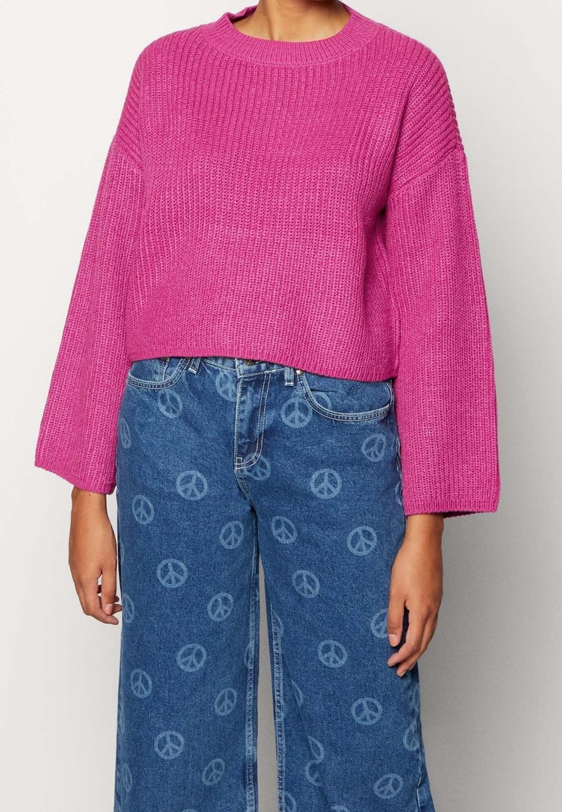 Rosa gestrickter Pullover mit weitem Ausschnitt und lockerer Passform; kombiniert mit blauen Jeans, die helle Friedenssymbol-Muster aufweisen.