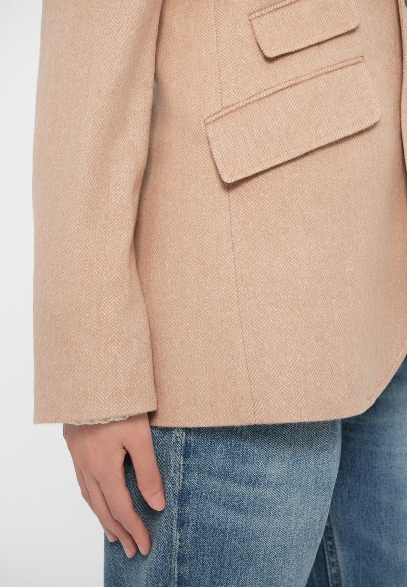 Blazer beige a spina di pesce con silhouette strutturata, due tasche frontali e una texture morbida. Abbinato a jeans denim azzurri.