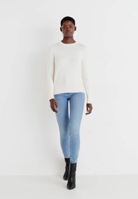 Top à manches longues crème, jean ajusté bleu clair et bottines noires. La texture lisse et le design moulant mettent en valeur une silhouette fluide.