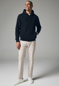Haut à capuche zippé noir avec une poche avant, associé à un pantalon beige clair et des baskets blanches. Le haut à capuche dispose d'une capuche avec cordon de serrage.