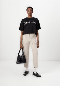 Black cropped t-shirt with white "Calvin Klein" logo, beige tailored pants, black handbag, black loafers, and white socks.