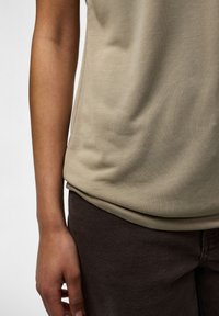 Gros plan d’une personne portant un haut beige et un pantalon marron foncé, montrant le bas du torse et le bras gauche détendu le long du corps.