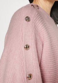 Maglione rosa lavorato a maglia con un motivo testurizzato e grandi bottoni in metallo dorato sulle spalle. Materiale morbido e caldo con una forma distintiva.